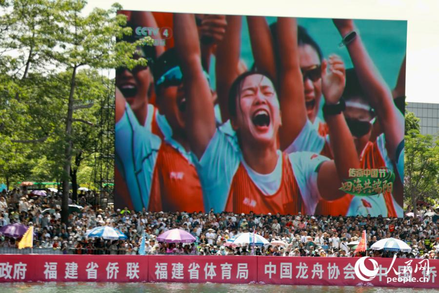赛场巨型大屏同步直播龙舟赛事画面，屏幕下方看台人头攒动。人民网 欧阳奕奕摄